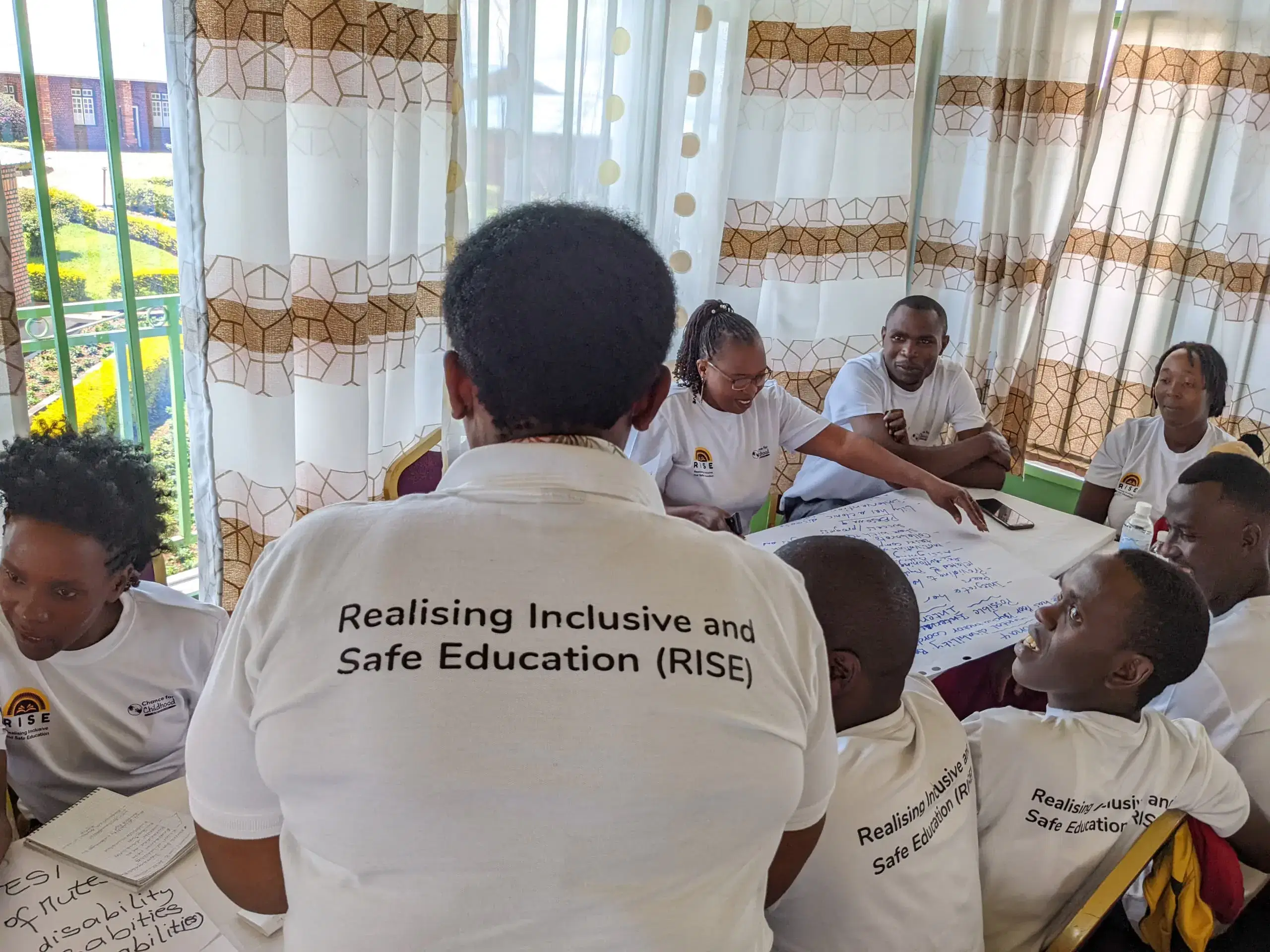 A man standing facing away from the camera. he is wearing a white top that reads Realising inclusive and safe education