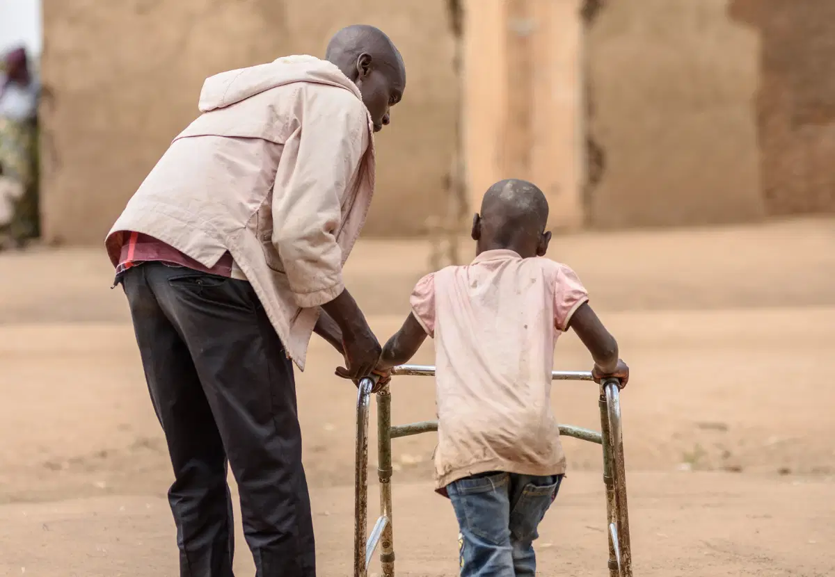 A man helps a young boy to walk using a walking frame.